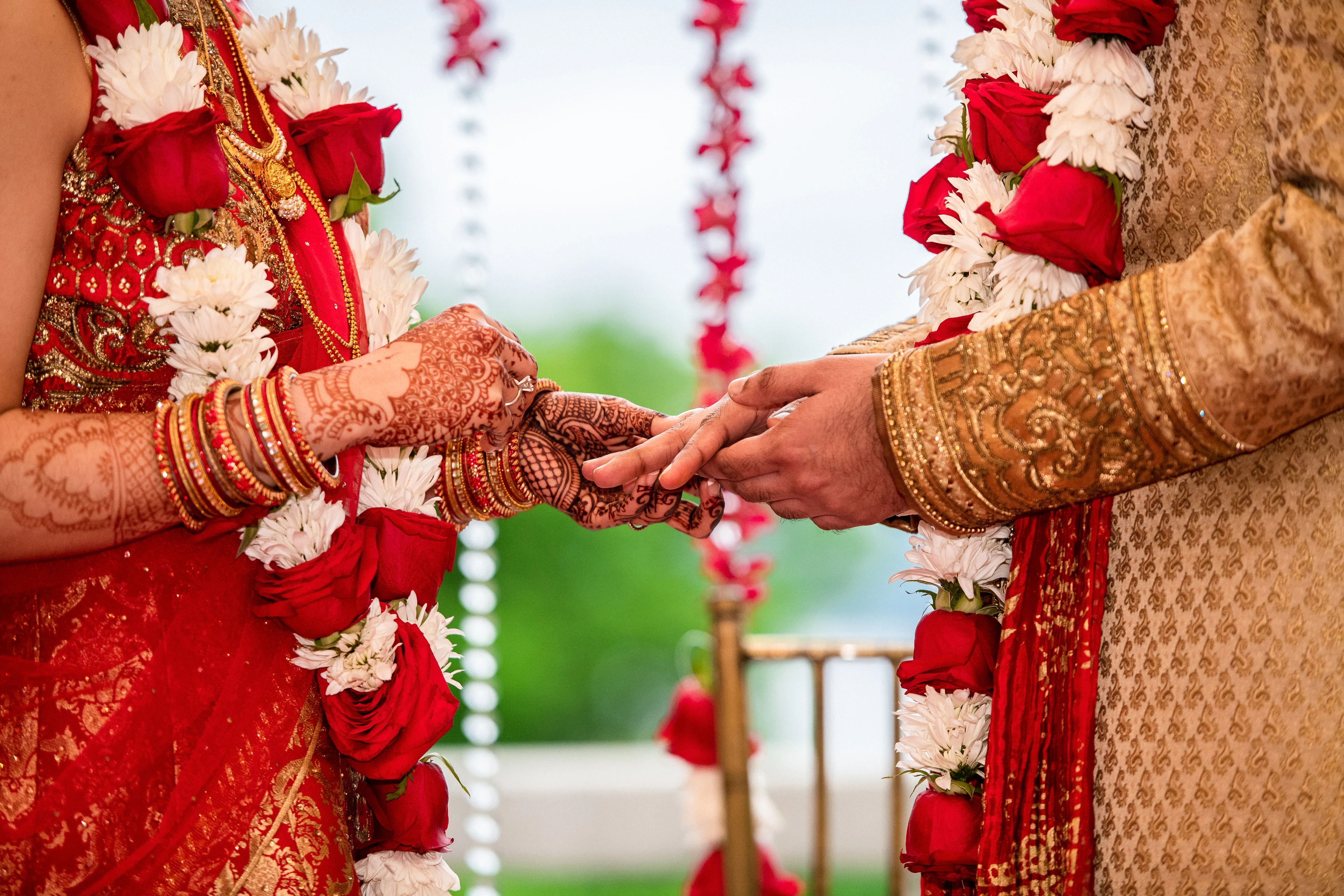 A bride and groom joining hennaed hands beneath rose and jasmine garlands at a Hindu wedding ceremony.