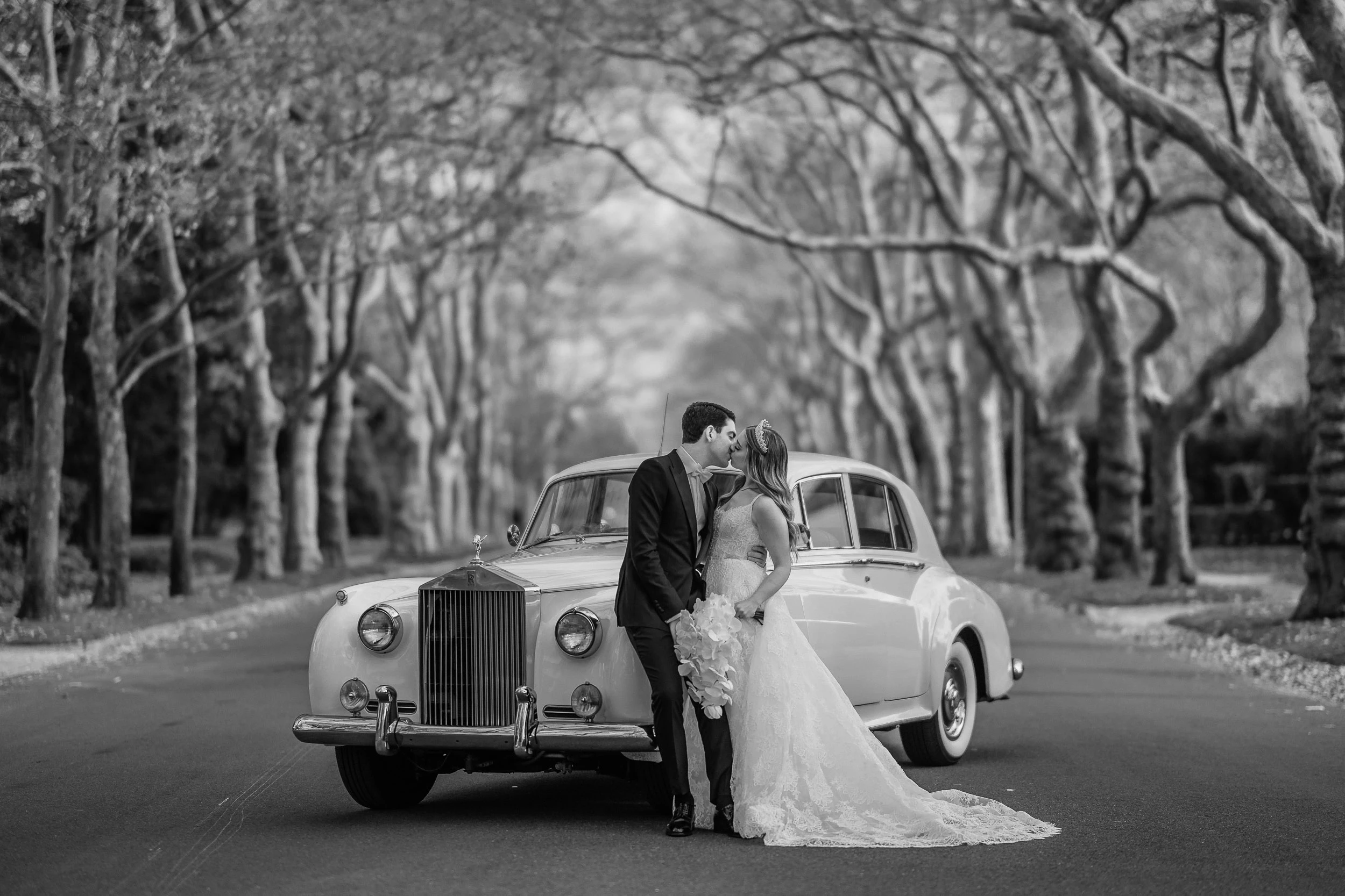 Black and white photograph of a couple kissing beside a vintage Rolls-Royce beneath an allée of bare plane trees.