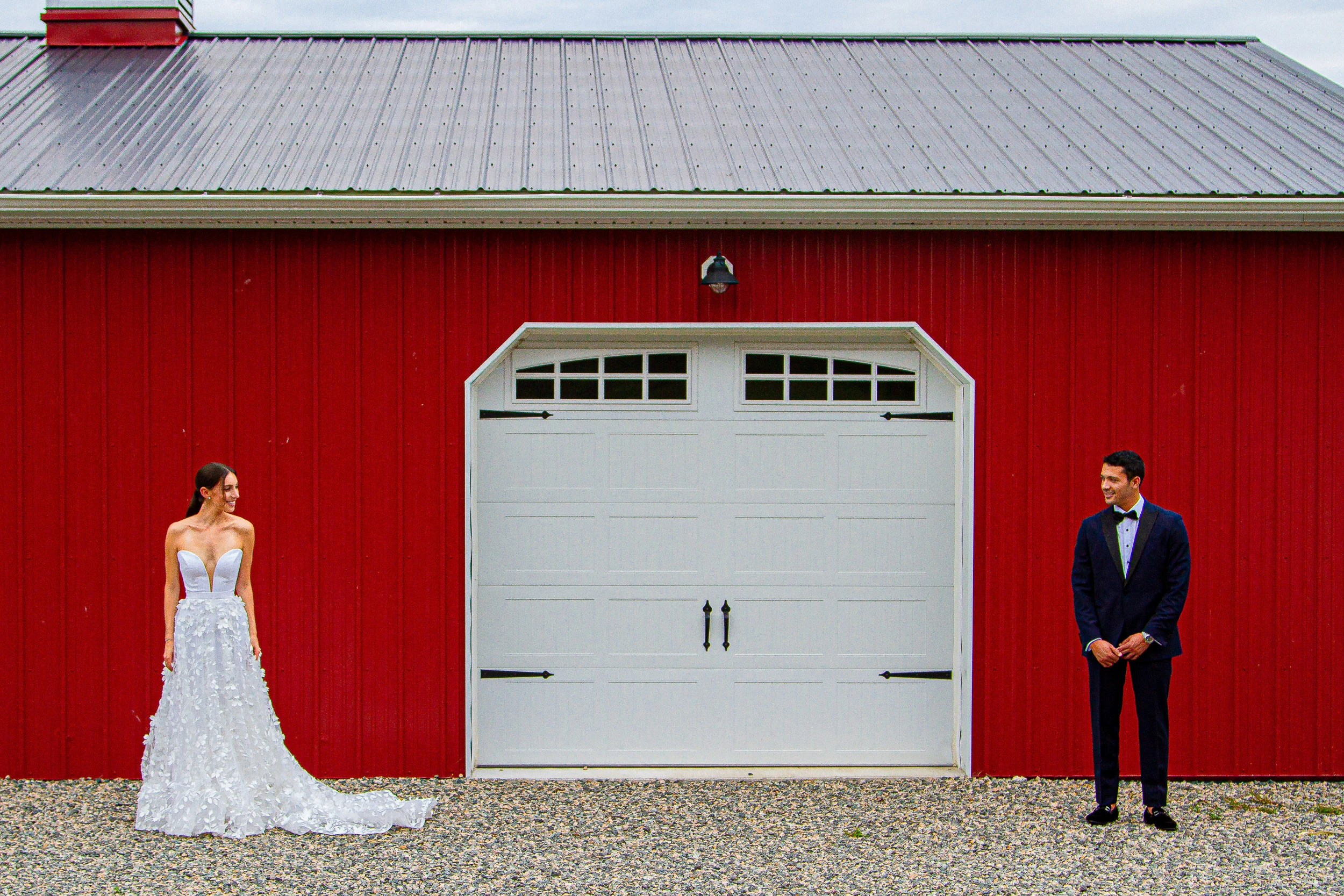 A bride and groom smiling at each other across the white door of a red barn before their first look.
